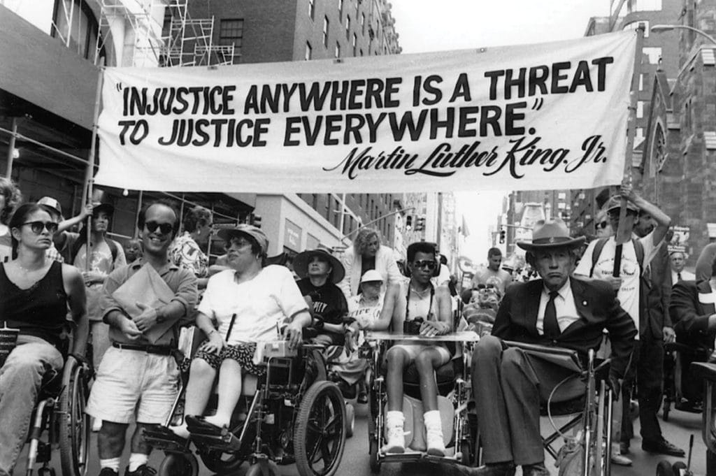 Injustice-anywhere Activists, including Judy Heumann and Justin Dart, at the Disability Independence March in Manhattan in July 1993, under a large banner of a Martin Luther King Jr. Quote: Injustice anywhere is a threat to justice everywhere.
