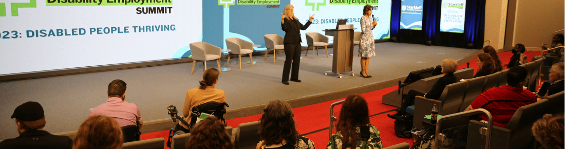 Auditorium stage with speaker and ASL interpreter the first couple rows of an engaged audience are visible.