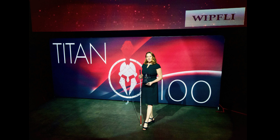 Lori Becker on a dramatically lit stage, holding her white cane and a glass award. The backdrop is red and black with a large Titan 100 logo, and Wipfli logo.