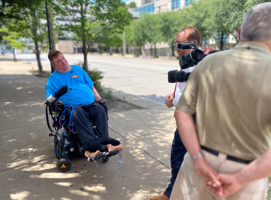 Steve seated in his power chair on a downtown sidewalk giving a TV news interview.