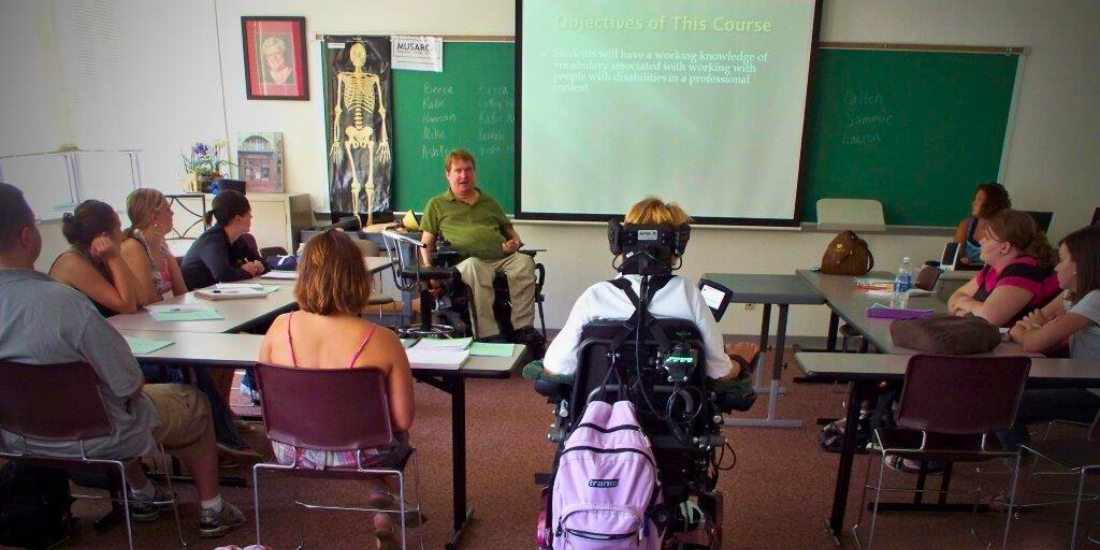 Steve, a power wheelchair user, teaches a college class. About 10 students are listening, seated at tables in a u-shape.