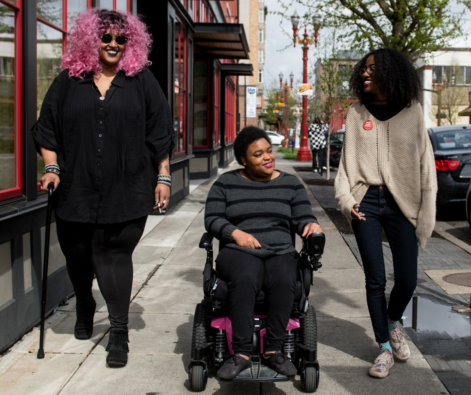 Three people strolling on the sidewalk in a vibrant city neighborhood. Two have apparent mobility disabilities. From Disabled and Here.