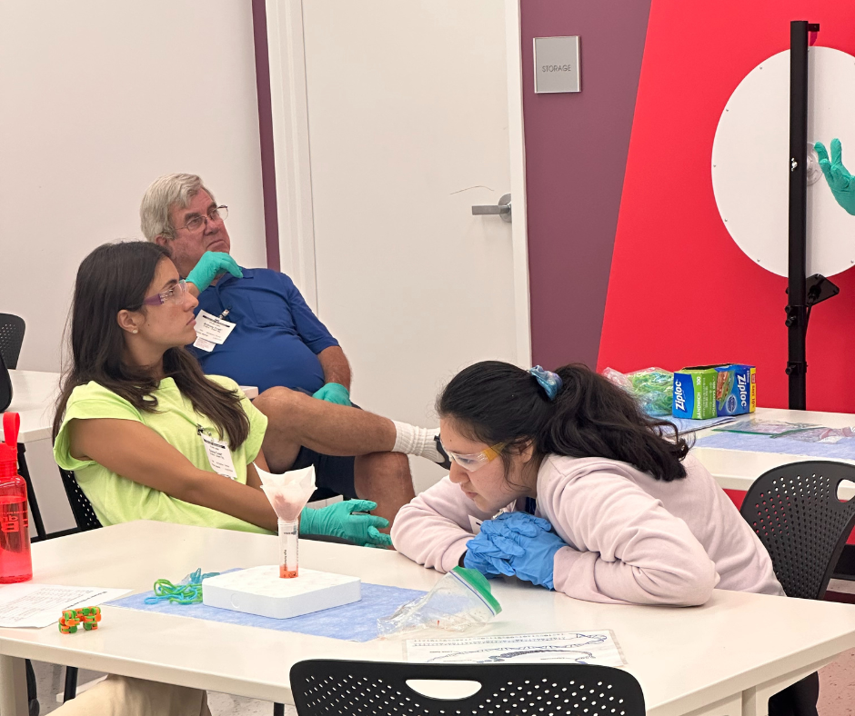 In a science learning lab, a student observes an experiment as Camp counselor and driver listen to an instructor.