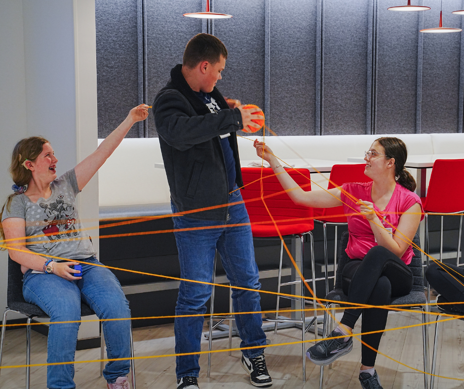 Three students laugh while holding parts of a large yarn web.